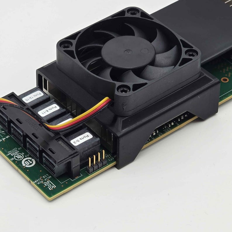 May include: Close-up of a black computer fan attached to a green circuit board. The fan has multiple blades and is mounted on a black housing. Several ports are labeled with white text. A multi-colored cable connects the fan to the board.