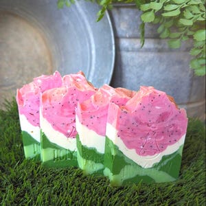 May include: Four rectangular bars of handmade soap arranged in a row. Each bar features a layered design resembling a watermelon slice, with a vibrant pink top section containing dark specks, a thin white middle layer, and a green base.