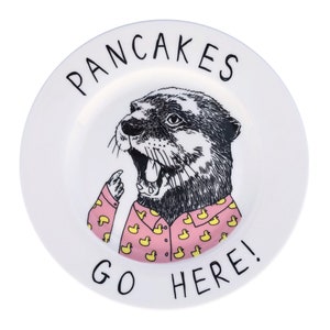 May include: White ceramic plate with a black and white illustration of an otter wearing a pink shirt with yellow rubber duckies. The text "PANCAKES GO HERE!" is written in black around the rim of the plate.