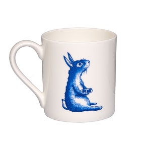 May include: White ceramic mug featuring a blue illustration of a rabbit holding a plate. The rabbit is looking to the right.