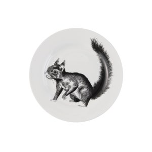 May include: A white ceramic plate featuring a black and white illustration of a squirrel. The squirrel is sitting on its haunches with its tail curled up behind it.