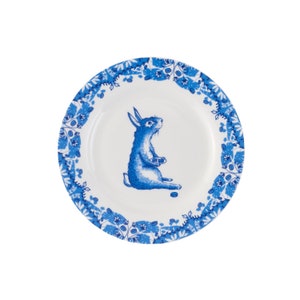 Puede incluir: Un plato de cer&aacute;mica blanco con un borde floral azul y blanco. Un conejo azul con la barriga blanca est&aacute; sentado en el centro del plato, sosteniendo un peque&ntilde;o objeto blanco.