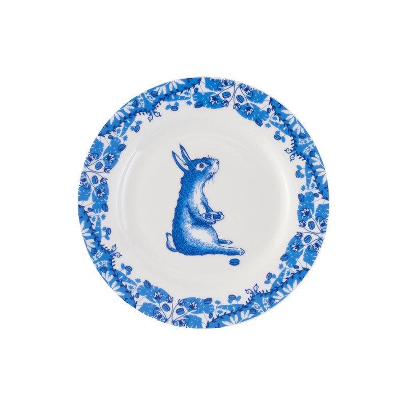 Puede incluir: Un plato de cer&aacute;mica blanco con un borde floral azul y blanco. Un conejo azul con la barriga blanca est&aacute; sentado en el centro del plato, sosteniendo un peque&ntilde;o objeto blanco.