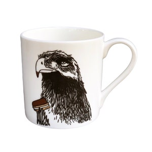 Può includere: Tazza in ceramica bianca con un'illustrazione in bianco e nero di una testa d'aquila con un biscotto marrone e bianco nel becco.