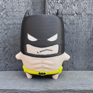 Peut inclure: Une petite figurine stylisée représentant un personnage portant une cagoule noire avec des découpes blanches pour les yeux, un torse beige aux muscles abdominaux définis et une ceinture utilitaire jaune vif avec un emblème noir.