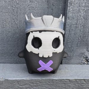 Peut inclure: Une petite figurine noire en trois dimensions en forme de créature arrondie. Elle présente un masque facial blanc en forme de crâne, une couronne argentée sur le dessus et une marque violette en forme de X sur la poitrine.