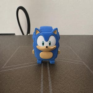 Puede incluir: Una figura impresa en 3D del personaje Sonic the Hedgehog, con cuerpo azul, hocico y vientre color canela, ojos blancos con iris verdes y una pequeña nariz negra. La figura tiene una textura en capas del proceso de impresión y se mantiene en pie.