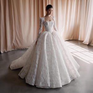 Könnte beinhalten: Ein weißes Brautkleid mit weitem Rock und langen Ärmeln. Das Kleid hat aufwendige Spitzendetails und einen Sweetheart-Ausschnitt. Das Kleid hat eine lange Schleppe und ein Oberteil im Bolero-Stil. Das Kleid ist bodenlang und scheint aus schimmerndem Stoff zu sein.