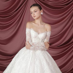 Peut inclure: Une robe de mari&eacute;e blanche &agrave; &eacute;paules d&eacute;nud&eacute;es avec de longues manches en dentelle et une jupe ample. Le corsage pr&eacute;sente des perles d&eacute;licates et un d&eacute;collet&eacute; en c&oelig;ur. La robe est en dentelle et en tulle, cr&eacute;ant un look romantique et &eacute;l&eacute;gant.