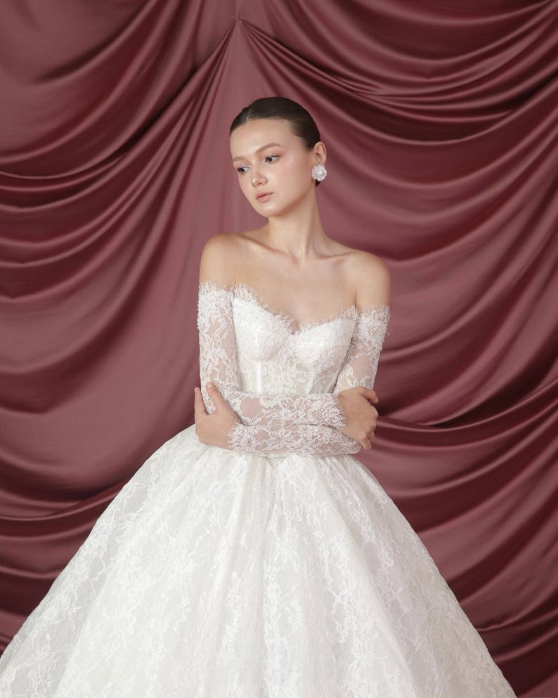 Peut inclure: Une robe de mari&eacute;e blanche &agrave; &eacute;paules d&eacute;nud&eacute;es avec de longues manches en dentelle et une jupe ample. Le corsage pr&eacute;sente des perles d&eacute;licates et un d&eacute;collet&eacute; en c&oelig;ur. La robe est en dentelle et en tulle, cr&eacute;ant un look romantique et &eacute;l&eacute;gant.