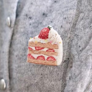 May include: A strawberry cake embroidered brooch featuring a slice of cake with layers of sponge, cream, and sliced strawberries, topped with a whole strawberry and cream. The text reads STRABERRY CAKE by EIGHT O'CLOCK STUDYHALL.