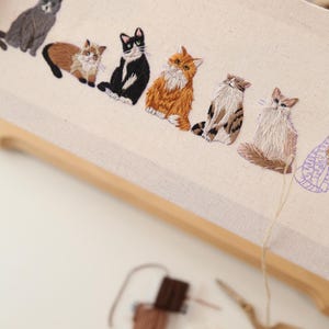 May include: Embroidered artwork featuring six different cat breeds in various colors, including gray, black, orange, and brown. The cats are stitched on a cream-colored fabric stretched within a wooden embroidery hoop. Embroidery floss and scissors are visible.