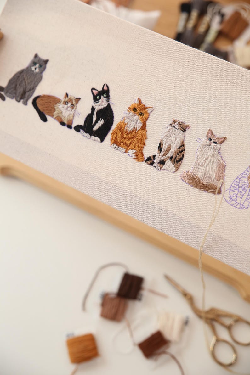 May include: Embroidered artwork featuring six different cat breeds in various colors, including gray, black, orange, and brown. The cats are stitched on a cream-colored fabric stretched within a wooden embroidery hoop. Embroidery floss and scissors are visible.