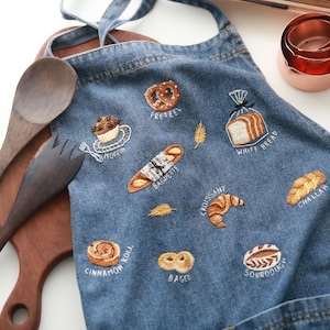 Peut inclure: Un tablier en jean bleu avec des images brodées de différents produits de boulangerie, notamment un bretzel, un muffin, une baguette, un croissant, un pain challah, un pain à la cannelle, un bagel et un pain au levain. Chaque image est étiquetée avec le nom du produit de boulangerie.
