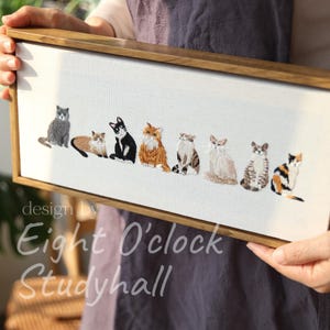 May include: Embroidered artwork featuring eight different cat breeds in various poses, set against a white background. The artwork is framed in a natural wood frame. The text "design by Eight O'clock Studyhall" is in the lower left corner.