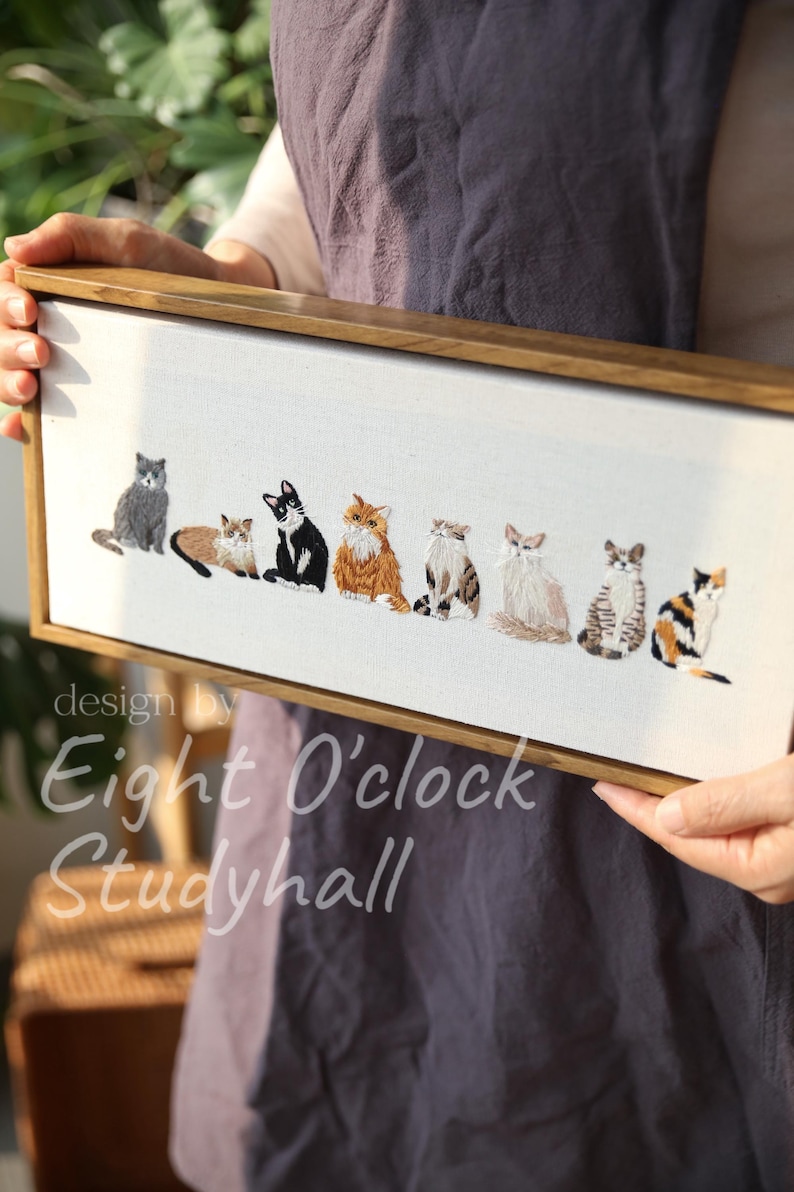 May include: Embroidered artwork featuring eight different cat breeds in various poses, set against a white background. The artwork is framed in a natural wood frame. The text "design by Eight O'clock Studyhall" is in the lower left corner.