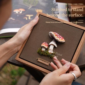 May include: A square wooden frame containing a brown fabric background with two embroidered Amanita muscaria mushrooms, green moss accents, and a small metal plaque labeled Amanita muscaria. The text reads Texture you can feel, Stitched to stand beyond the surface, Raised forms and layered details create true dimension, and O'clock Studyshell.
