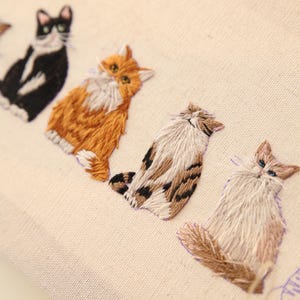 May include: Embroidered fabric featuring a row of stylized cat portraits. The cats are rendered in various colors, including black and white, orange, and brown, with detailed stitching. The background is a light beige.