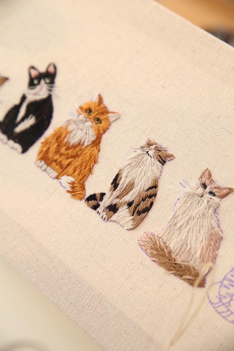 May include: Embroidered fabric featuring a row of stylized cat portraits. The cats are rendered in various colors, including black and white, orange, and brown, with detailed stitching. The background is a light beige.