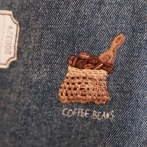Puede incluir: Dise&ntilde;o bordado en tela vaquera de un saco de arpillera marr&oacute;n lleno de granos de caf&eacute;, con el texto "COFFEE BEANS" cosido debajo.