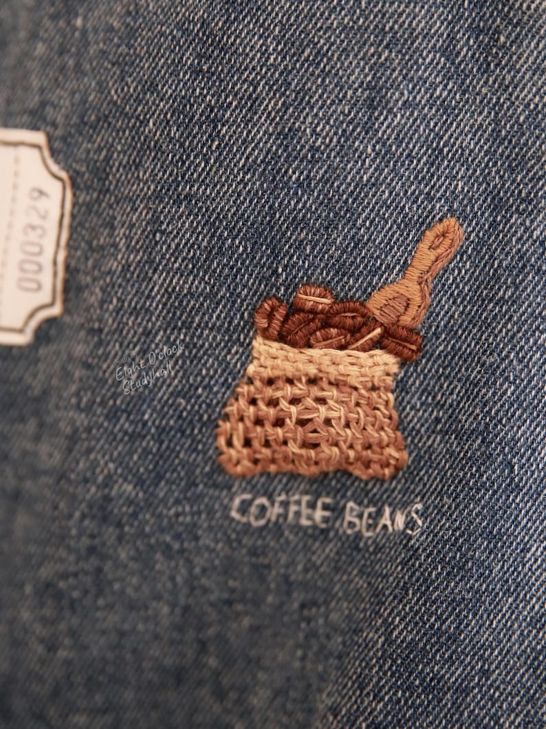 Puede incluir: Dise&ntilde;o bordado en tela vaquera de un saco de arpillera marr&oacute;n lleno de granos de caf&eacute;, con el texto "COFFEE BEANS" cosido debajo.