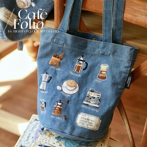 Puede incluir: Una bolsa de lona azul con dise&ntilde;os bordados con tem&aacute;tica de caf&eacute;, que incluyen granos de caf&eacute;, una prensa francesa, un Chemex, una cafetera italiana, un capuchino, una m&aacute;quina de espresso, un molinillo de mano y un boleto que dice "Ticket to Coffee Land".