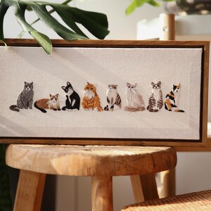 May include: Embroidered artwork featuring seven different cat breeds in various colors, set against a neutral canvas. The cats are depicted in a row, each with unique markings and poses, framed in a natural wood. A charming piece for cat lovers.