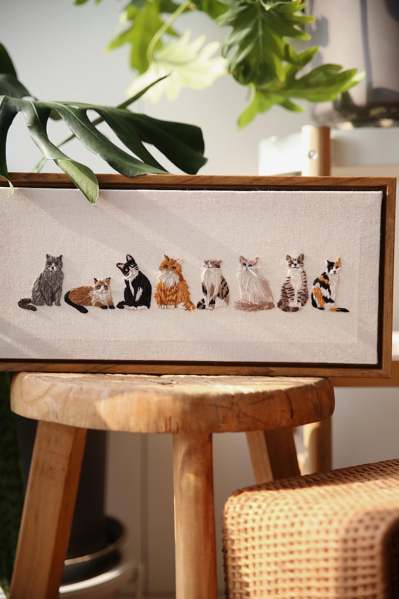 May include: Embroidered artwork featuring seven different cat breeds in various colors, set against a neutral canvas. The cats are depicted in a row, each with unique markings and poses, framed in a natural wood. A charming piece for cat lovers.