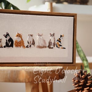 May include: Embroidered artwork featuring a row of six cats in various poses and colors, set against a neutral background. The artwork is framed in a wooden frame. The text "design by Eight O'clock Studyhall" is visible.