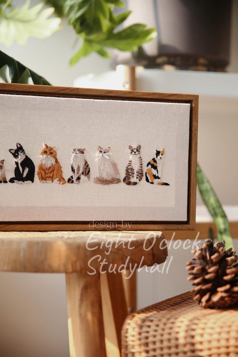 May include: Embroidered artwork featuring a row of six cats in various poses and colors, set against a neutral background. The artwork is framed in a wooden frame. The text "design by Eight O'clock Studyhall" is visible.