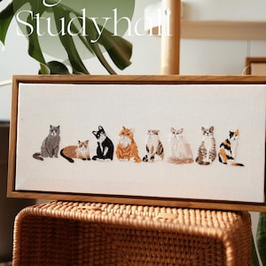 May include: Embroidered artwork featuring a row of seven cats in various colors and patterns, set against a white canvas. The artwork is framed in a natural wood frame. The text "design by Eight O'clock Studyhall" is at the top.