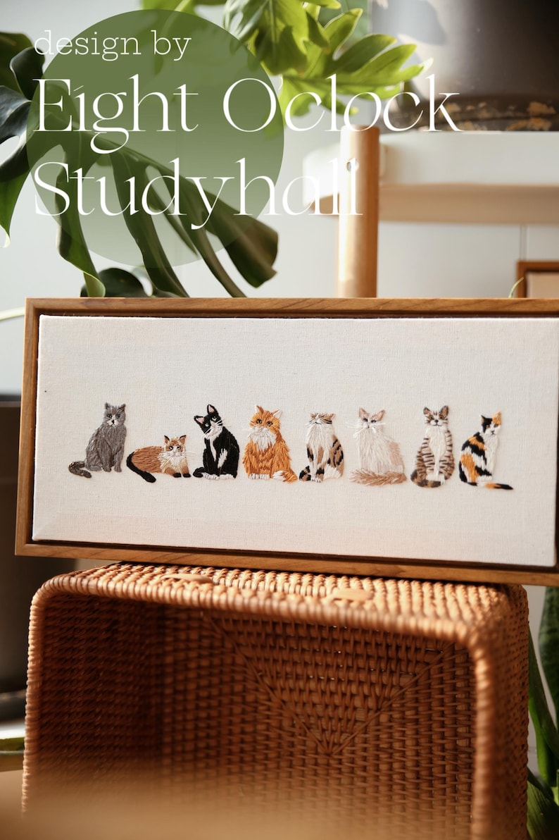 May include: Embroidered artwork featuring a row of seven cats in various colors and patterns, set against a white canvas. The artwork is framed in a natural wood frame. The text "design by Eight O'clock Studyhall" is at the top.