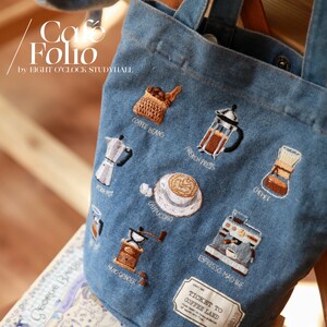 Puede incluir: Una bolsa de lona azul con ilustraciones bordadas con tem&aacute;tica de caf&eacute;. La bolsa presenta im&aacute;genes de granos de caf&eacute;, una prensa francesa, una cafetera moka, un capuchino, un molinillo de mano, una m&aacute;quina de espresso y un filtro de caf&eacute;. El texto "Cafe Folio by EIGHT O'CLOCK STUDYHALL" est&aacute; impreso en la bolsa. La bolsa tambi&eacute;n tiene una etiqueta que dice "TICKET TO COFFEE LAND".