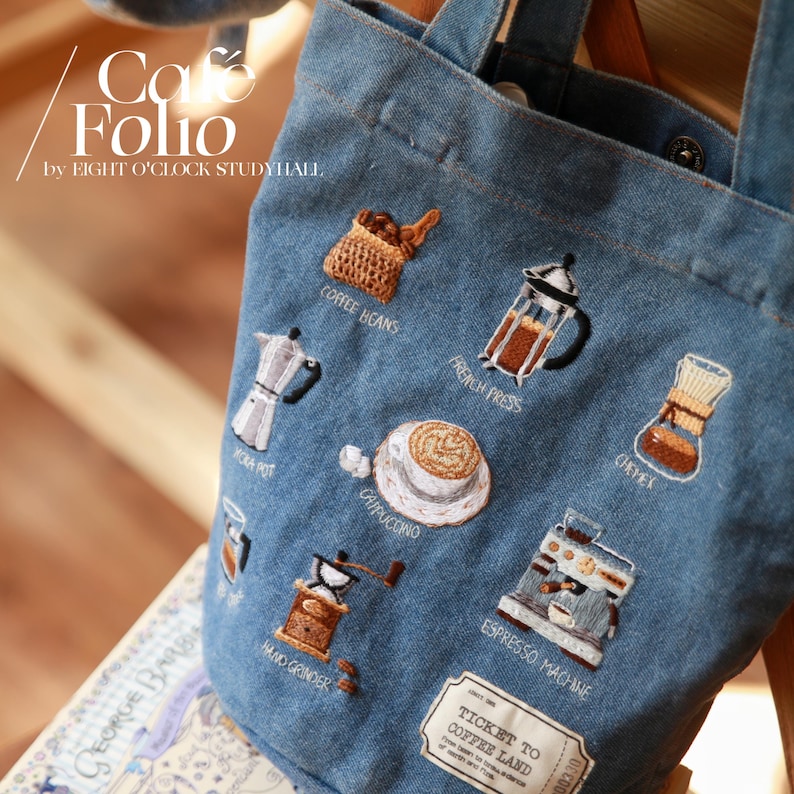 Puede incluir: Una bolsa de lona azul con ilustraciones bordadas con tem&aacute;tica de caf&eacute;. La bolsa presenta im&aacute;genes de granos de caf&eacute;, una prensa francesa, una cafetera moka, un capuchino, un molinillo de mano, una m&aacute;quina de espresso y un filtro de caf&eacute;. El texto "Cafe Folio by EIGHT O'CLOCK STUDYHALL" est&aacute; impreso en la bolsa. La bolsa tambi&eacute;n tiene una etiqueta que dice "TICKET TO COFFEE LAND".