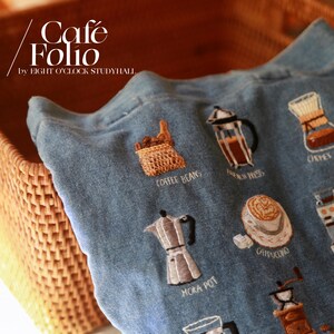 Puede incluir: Una bolsa de lona azul con ilustraciones bordadas con tem&aacute;tica de caf&eacute;, incluyendo granos de caf&eacute;, una prensa francesa, una cafetera italiana y un capuchino. El texto "Cafe Folio by Eight O'Clock Studyhall" est&aacute; bordado en la bolsa.