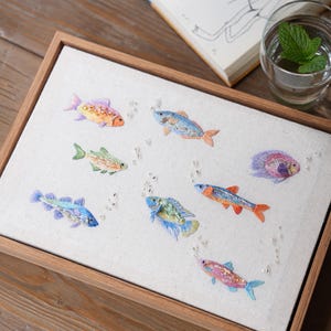 May include: A rectangular wooden frame containing a white fabric panel embroidered with eight colourful fish. Each fish features intricate thread work and small beads, with scattered clear beads representing bubbles floating between the fish figures.