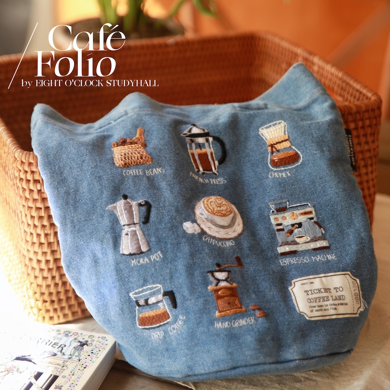 Puede incluir: Una bolsa de lona azul con ilustraciones bordadas con tem&aacute;tica de caf&eacute;, que incluyen granos de caf&eacute;, una prensa francesa, un Chemex, una cafetera Moka, un capuchino, una m&aacute;quina de espresso, caf&eacute; de goteo y un molinillo de mano. La bolsa tiene el texto "Cafe Folio by Eight O'Clock Studyhall" y "Ticket to Coffee Land" impreso en ella.