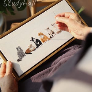 May include: Embroidered artwork featuring six stylized cats in various colors, including gray, brown, black, and orange. The design is framed in a wooden rectangle. The text "design by Eight O'clock Studyhall" is in the upper left corner.
