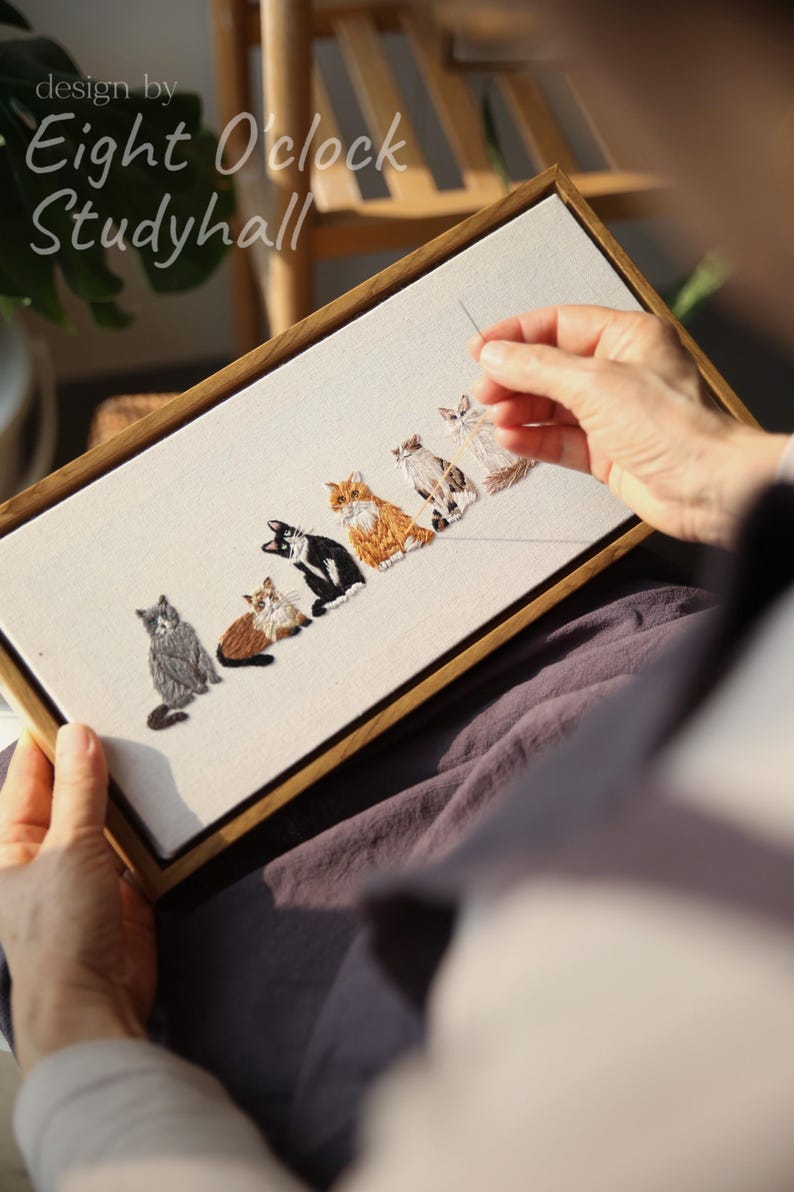 May include: Embroidered artwork featuring six stylized cats in various colors, including gray, brown, black, and orange. The design is framed in a wooden rectangle. The text "design by Eight O'clock Studyhall" is in the upper left corner.