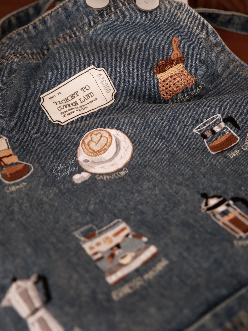Puede incluir: Un delantal de mezclilla con dise&ntilde;os bordados con tem&aacute;tica de caf&eacute;, que incluyen un boleto a la tierra del caf&eacute;, una bolsa de granos de caf&eacute;, un capuchino, una cafetera de goteo, una m&aacute;quina de espresso y una cafetera italiana. El texto en el boleto dice "Ticket to Coffee Land" y "From dawn to sunrise a chance of sweet, warm rain."