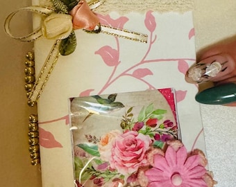 Puede incluir: Un pequeño diario hecho a mano con un diseño floral. La portada está decorada con encaje, una flor rosa y una cinta dorada con un lazo. El diario tiene una encuadernación con cuentas doradas.