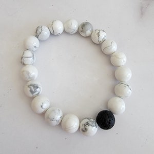 Puede incluir: Una pulsera de cuentas blancas con vetas grises y una sola cuenta de lava negra. Las cuentas redondas están ensartadas, creando un accesorio simple y elegante, adecuado para diversas ocasiones.