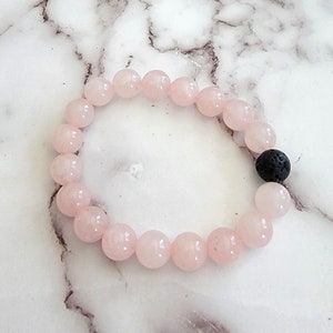 Puede incluir: Un brazalete de cuentas de cuarzo rosa con una cuenta de lava negra. El brazalete está hecho de piedras naturales y está diseñado para ser usado en la muñeca.