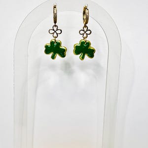 May include: Gold-coloured hoop earrings featuring a green and gold shamrock charm. The earrings are displayed on a clear acrylic stand against a white backdrop. These earrings are a festive accessory, perfect for St. Patrick's Day.