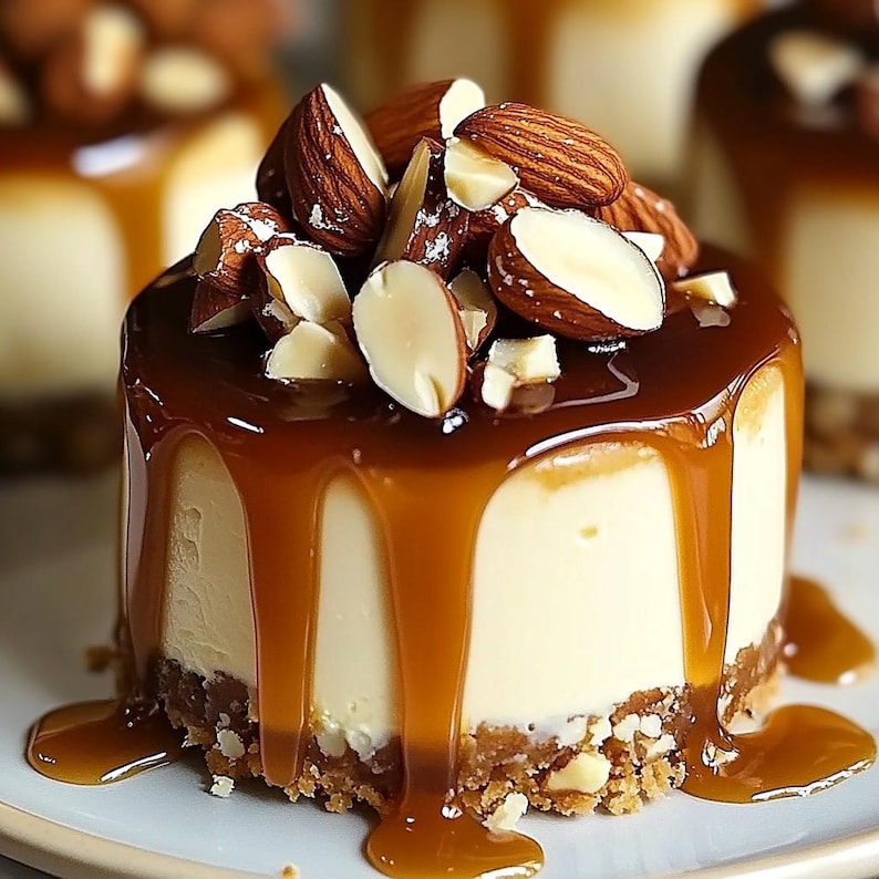 May include: A small cheesecake topped with caramel sauce and sliced almonds. The cheesecake is on a white plate with a small amount of caramel sauce around it.