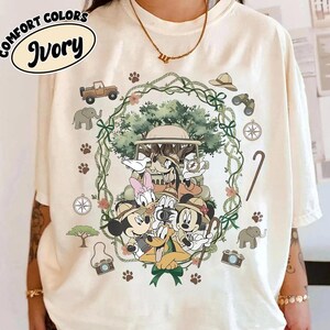 Puede incluir: Camiseta Comfort Colors color marfil con un dise&ntilde;o de tem&aacute;tica safari. El gr&aacute;fico presenta a Mickey Mouse, el Pato Donald y sus amigos, enmarcados por una corona verde con im&aacute;genes de safari como un jeep, una br&uacute;jula y unos prism&aacute;ticos. La camiseta tiene el texto "Comfort Colors" e "Ivory".