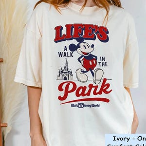Puede incluir: Camiseta de color marfil con un gr&aacute;fico de Mickey Mouse y el texto "LIFE'S A WALK IN THE PARK" en rojo y azul. El dise&ntilde;o incluye un castillo y las palabras "Walt Disney World".