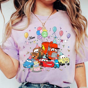May include: A light purple t-shirt featuring a colorful graphic of animated cars, balloons, and a birthday cake. The design includes the text "Name" and "2". The shirt is a short-sleeved crew neck.