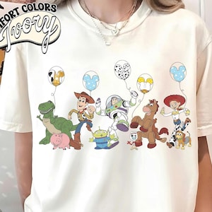 K&ouml;nnte beinhalten: Cremefarbenes T-Shirt mit einer bunten Grafik von Zeichentrickfiguren, die Luftballons halten. Die Figuren umfassen einen gr&uuml;nen Dinosaurier, ein Schwein, einen Cowboy, einen Astronauten, einen Au&szlig;erirdischen, ein Pferd und ein Cowgirl. Das Shirt hat den Schriftzug "Comfort Colors Ivory".