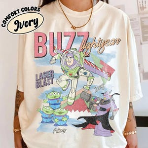 Puede incluir: Camiseta de color marfil con un gr&aacute;fico de Buzz Lightyear, extraterrestres y Zurg de la pel&iacute;cula Toy Story. El dise&ntilde;o incluye las palabras "BUZZ lightyear" y "LASER BLAST" en rosa y azul. La camiseta es de la marca Comfort Colors.